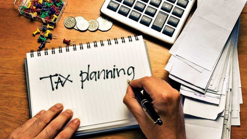 A hand with a pen that has written "tax planning" on a notepad surrounded by other desk objects.